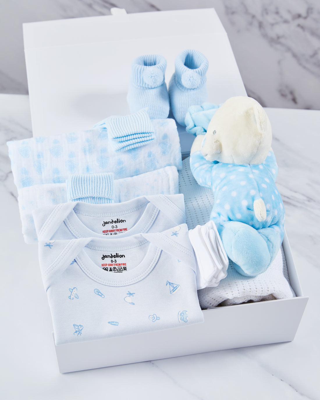 Baby boy clothing gift set, inside a white magnetic box, packed with 2 baby vests, 2 muslins, 2 pairs of socks, acrylic booties, 2 pairs of mittens, cellular blanket and blue polka dot teddy bear toy. 